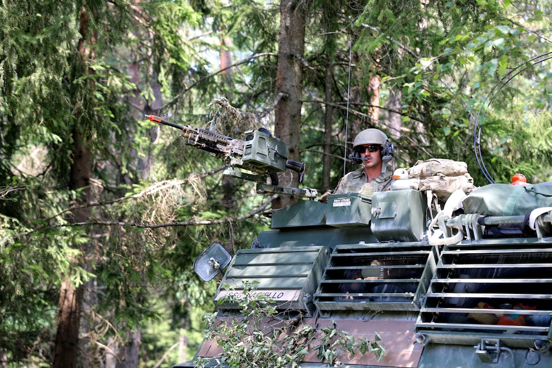 A soldier looks out from the top of tank in a forested area.