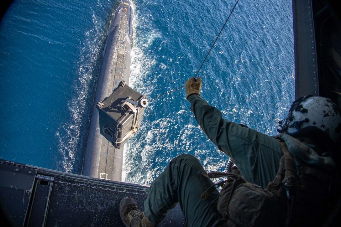 A Marine holds a cable lowering a box of supplies from an aircraft.