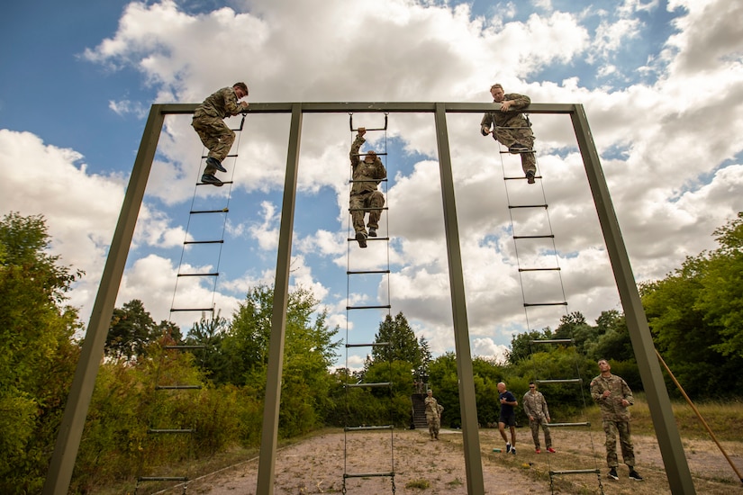 Three soldiers climb ladders next to each other during a competition as fellow soldiers watch from below.