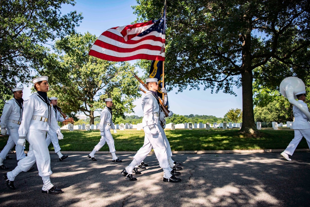 Service members march carrying weapons during a funeral escort at a cemetery.