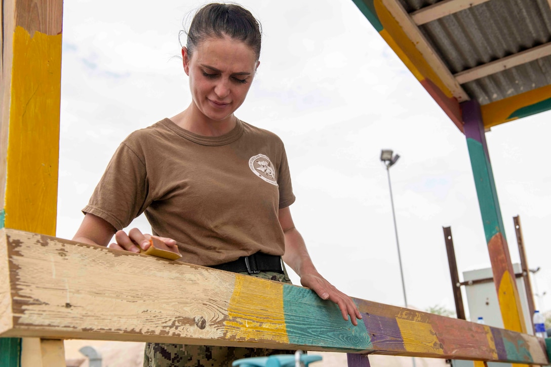 A sailor uses a tool to sand a wood bench.