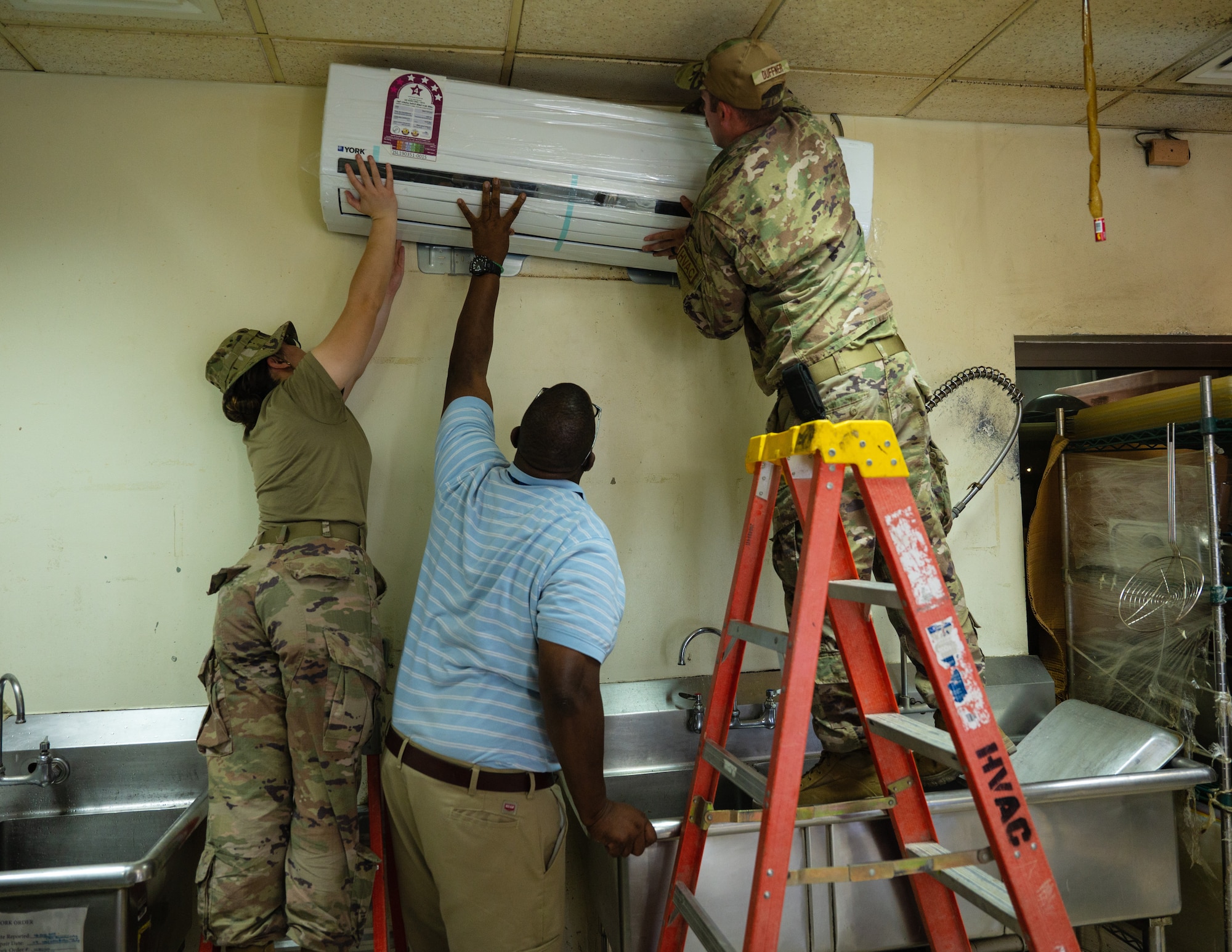 U.S. Air Force Airman 1st Class Haunanionapua Wright (left) and U.S. Air Force Staff Sgt. Jonathan Duffner (right), heating ventilation and air conditioning airman from the 379th Expeditionary Civil Engineering Squadron, work with Alternate Dining Facility Manager Shenan Goff (Center) with hanging a mini-split air conditioner in the Hot Grab, July 29, 2022 at Al Udeid Air Base, Qatar. Once properly hung on the wall bracket, the unit was wired and connected to the existing pipes making it ready for use. (U.S. Air Force photo by Staff Sgt. Dana Tourtellotte)