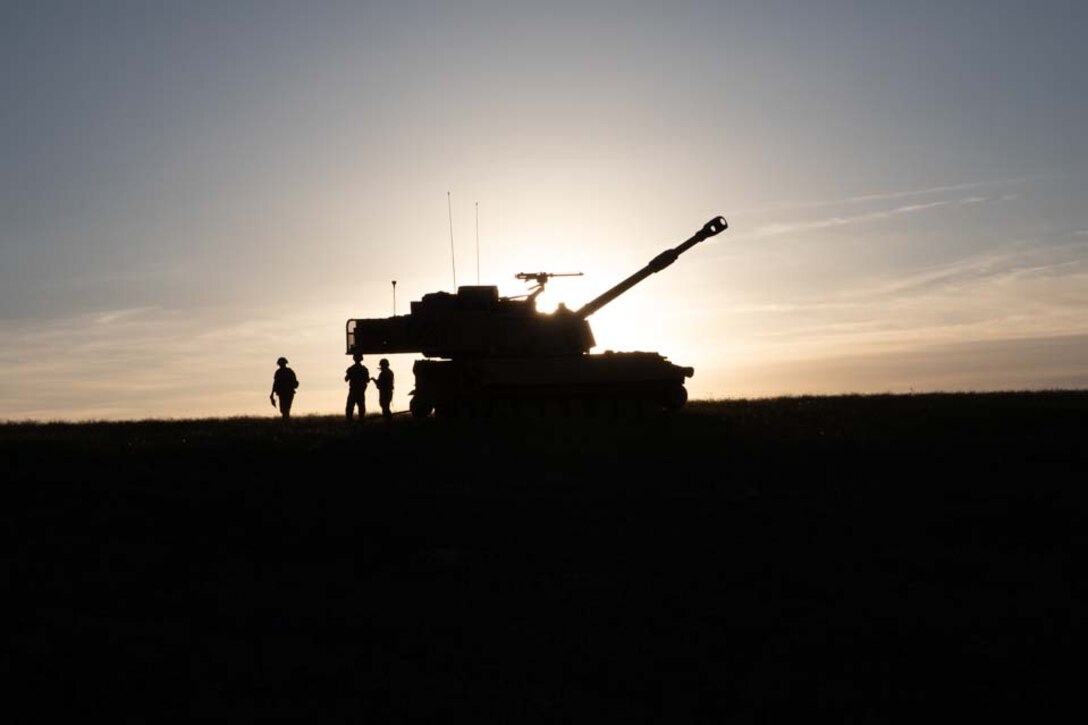 A howitzer and a group of soldiers are silhouetted against the sun.