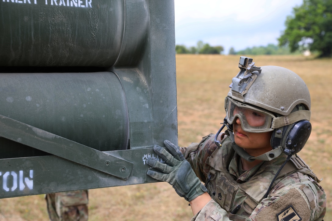 A soldier wearing a helmet and gloves pushes on a large piece of weaponry.