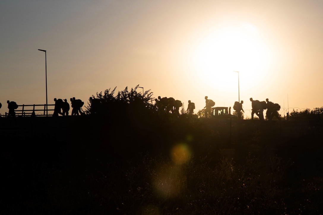 Soldiers march through a field at sunset with large ruck sacks on their backs.