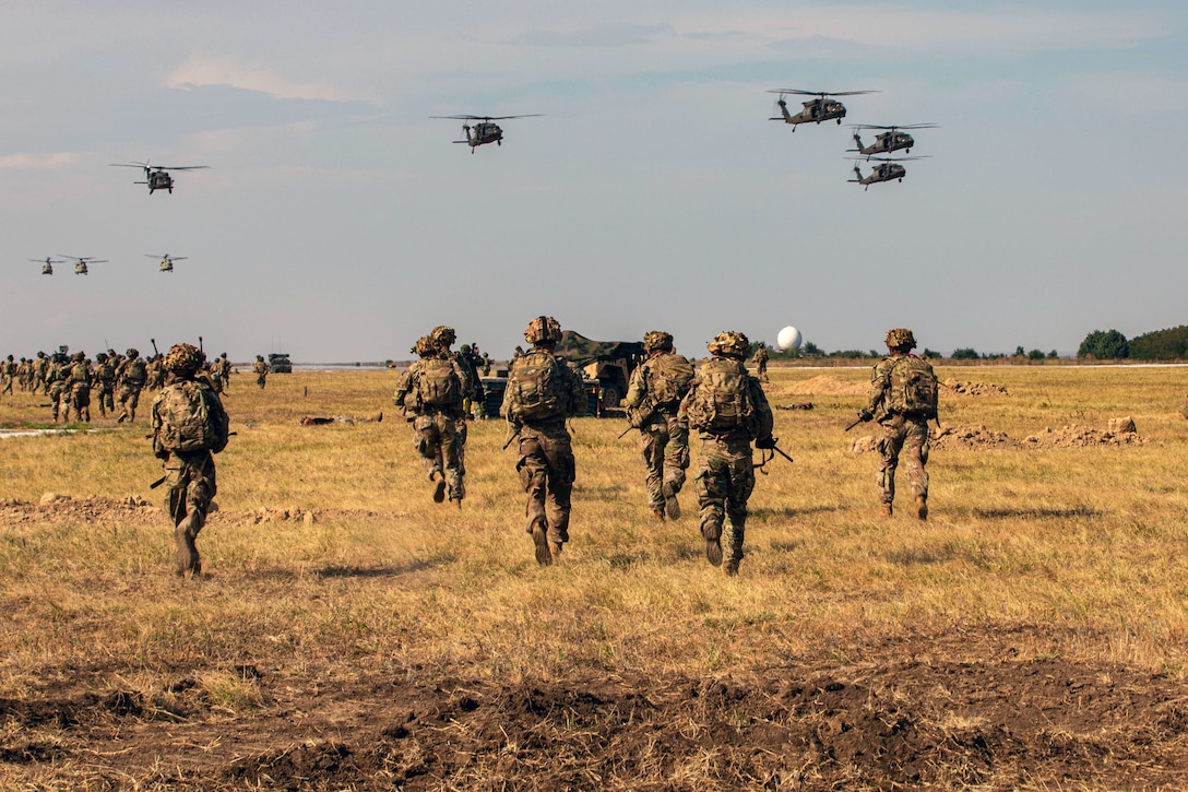 Many armed soldiers walk through a field as military helicopters fly overhead.
