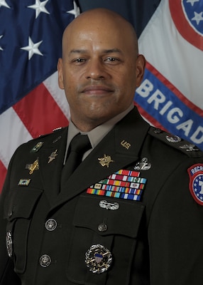 man wearing U.S. Army uniform standing in front of two flags.