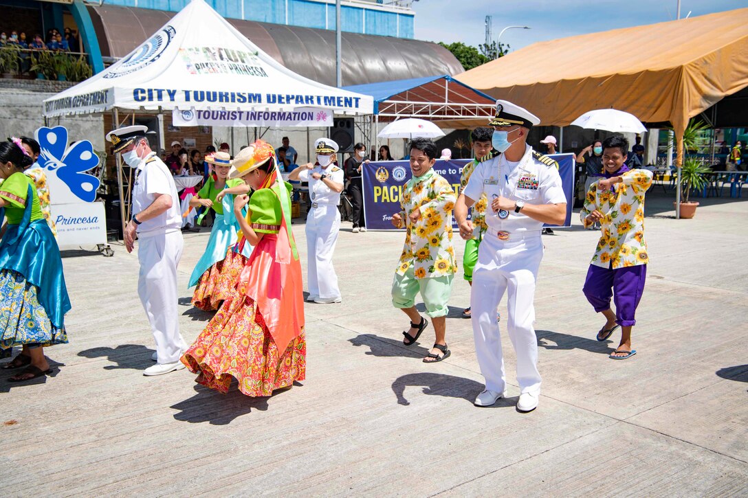 Three sailors dance with a colorful group at a pier with tents in the background.