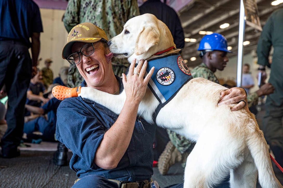 A dog wearing orange booties licks a smiling sailor's face, displacing her glasses as she sits on a ship's floor.