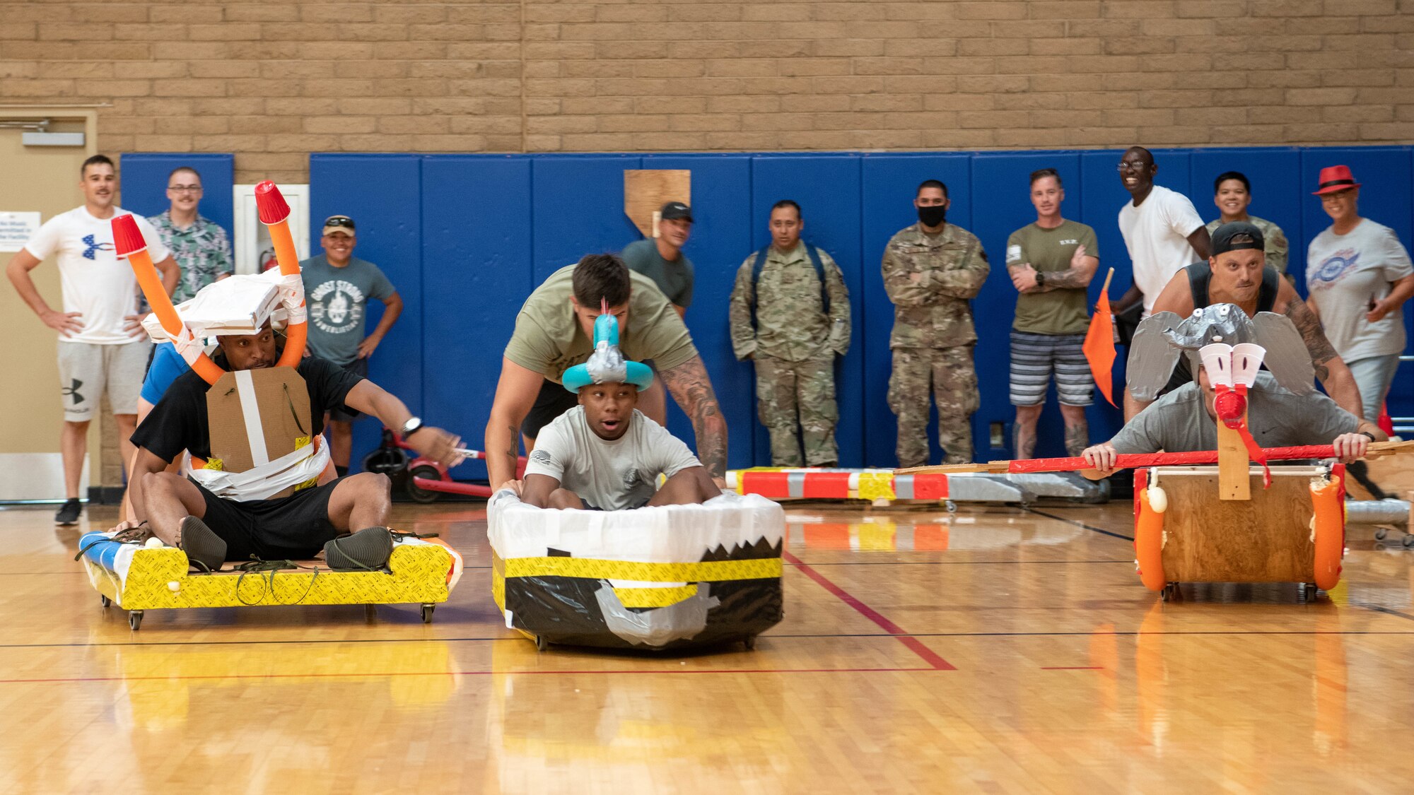 Airman line up on a starting line with their homemade vehicles during a race inside of a gym.