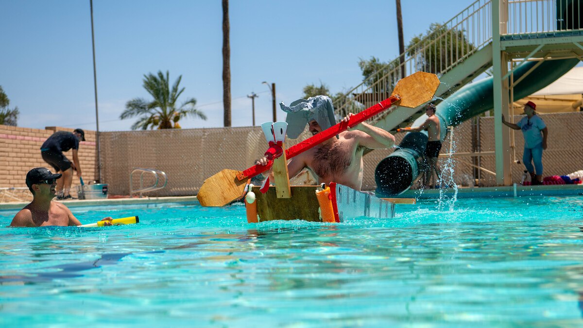 An Airman uses a makeshift paddle to steer a homemade boat in a pool.