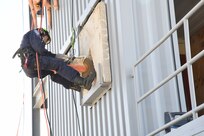 Rescue worker cuts through a concrete wall while he is suspended by rappelling ropes.