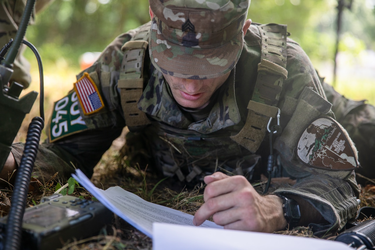 A soldier lies in the forest and reads paperwork by a portable radio.