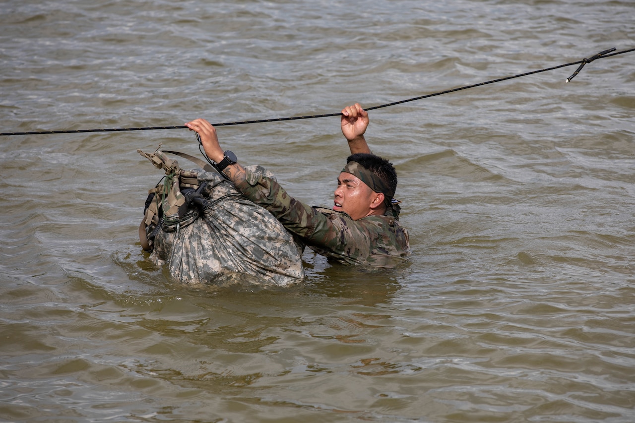 A soldier holds onto a rope over his head while navigating through deep water with a large ruck.