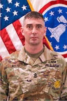 Soldier in uniform in front of flag.