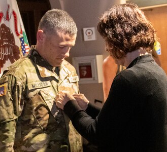 Jeanie Greenwalt, mother of newly promoted Chief Warrant Officer 4 Tim Milligan, of Illiopolis, Illinois, Warrant Officer Strength Manager, Recruiting and Retention Battalion, Illinois Army National Guard, secures new rank on his uniform during a promotion ceremony Aug. 3 at the Illinois State Military Museum, Springfield, Illinois.