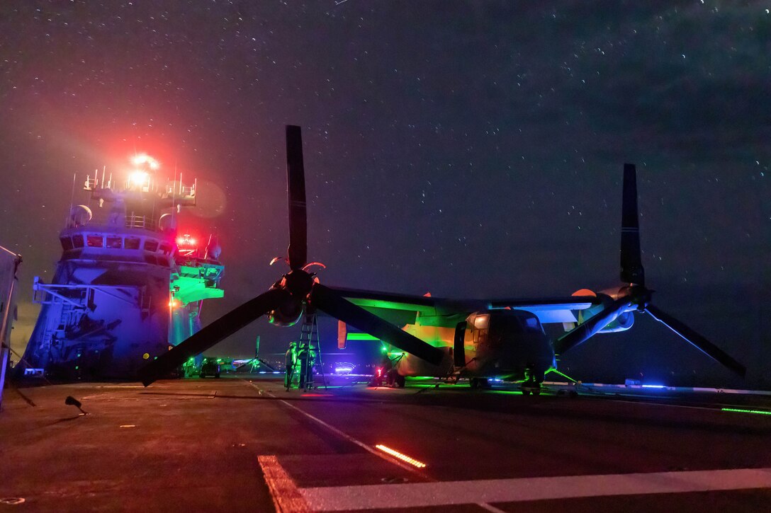 Service members work on an aircraft aboard a ship at sea illuminated by colorful lights.