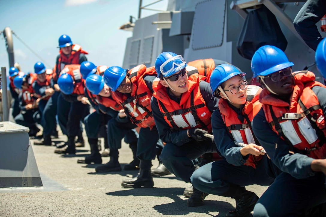 Sailors wearing hard hats and flotation devices stand in line pulling a rope aboard a ship at sea.