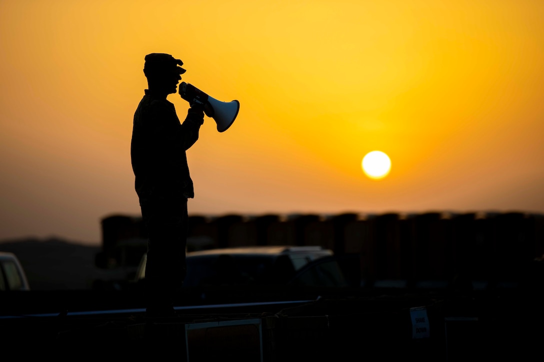 A soldier shown in silhouette speaks into a megaphone as the sun shines behind.