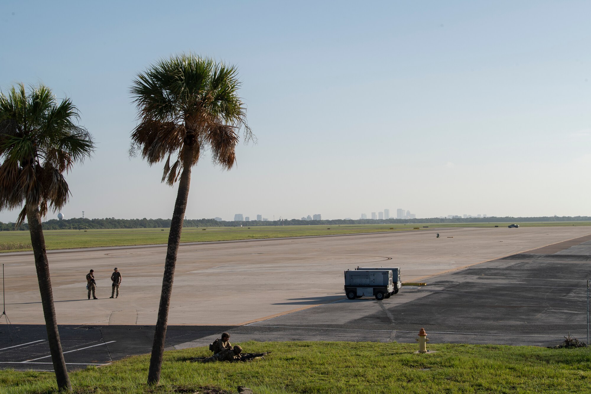A photo of a flight line with a set of sparse palm trees to the left framing two airmen that are standing in the middle of them on the flight line.