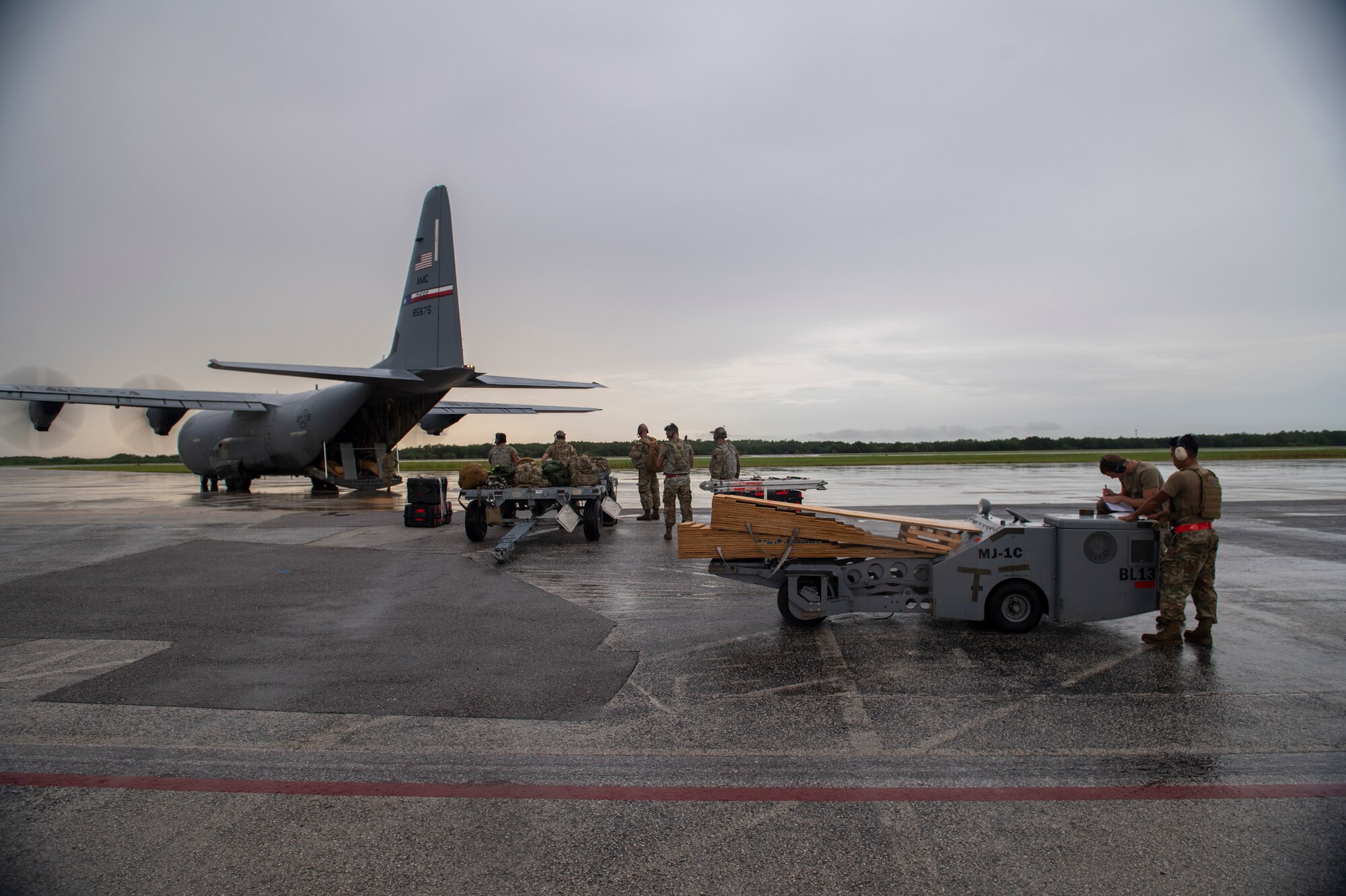 A photo of an aircraft in the back ground of the photo, a grey/yellow sunset, with airmen and weapons load equipment in the foreground.