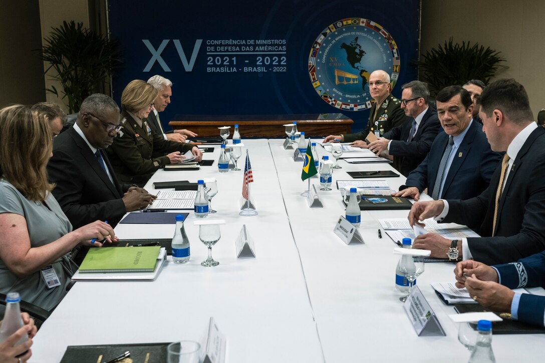 A group of people sit at a table, with the left side sitting behind a small U.S. flag and the right site sitting behind a small Brazilian flag. The wall in the background displays a logo for the 15th Conference of Defense Ministers of the Americas in Brazil.