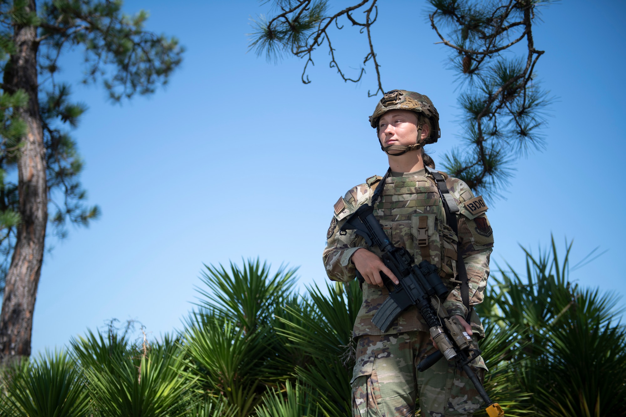 A photo of an Airman standing with her M-4 Carbine pointed towards the ground, green sprouting palm trees casting behind her on the ground with a bright blue sky.