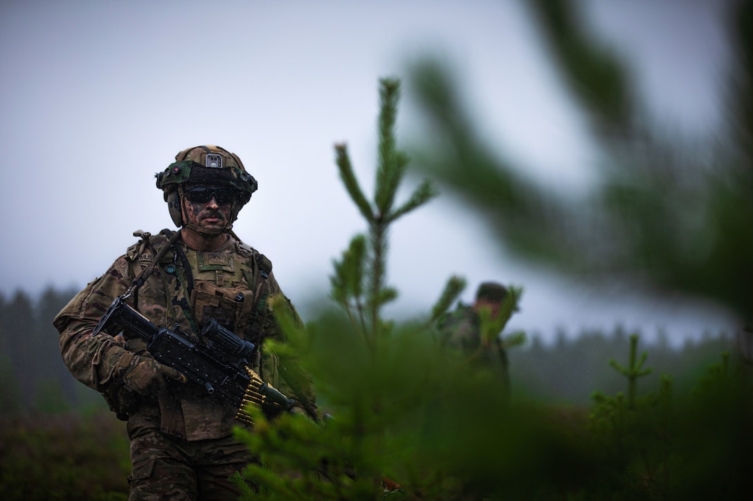 A soldier in camouflage holds a weapon as he walks through grove of head-high pine trees; another soldier is in the background.