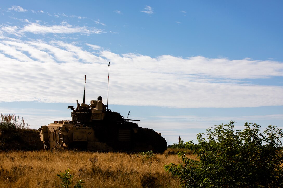 A soldier rides in a tank across a grassy area.