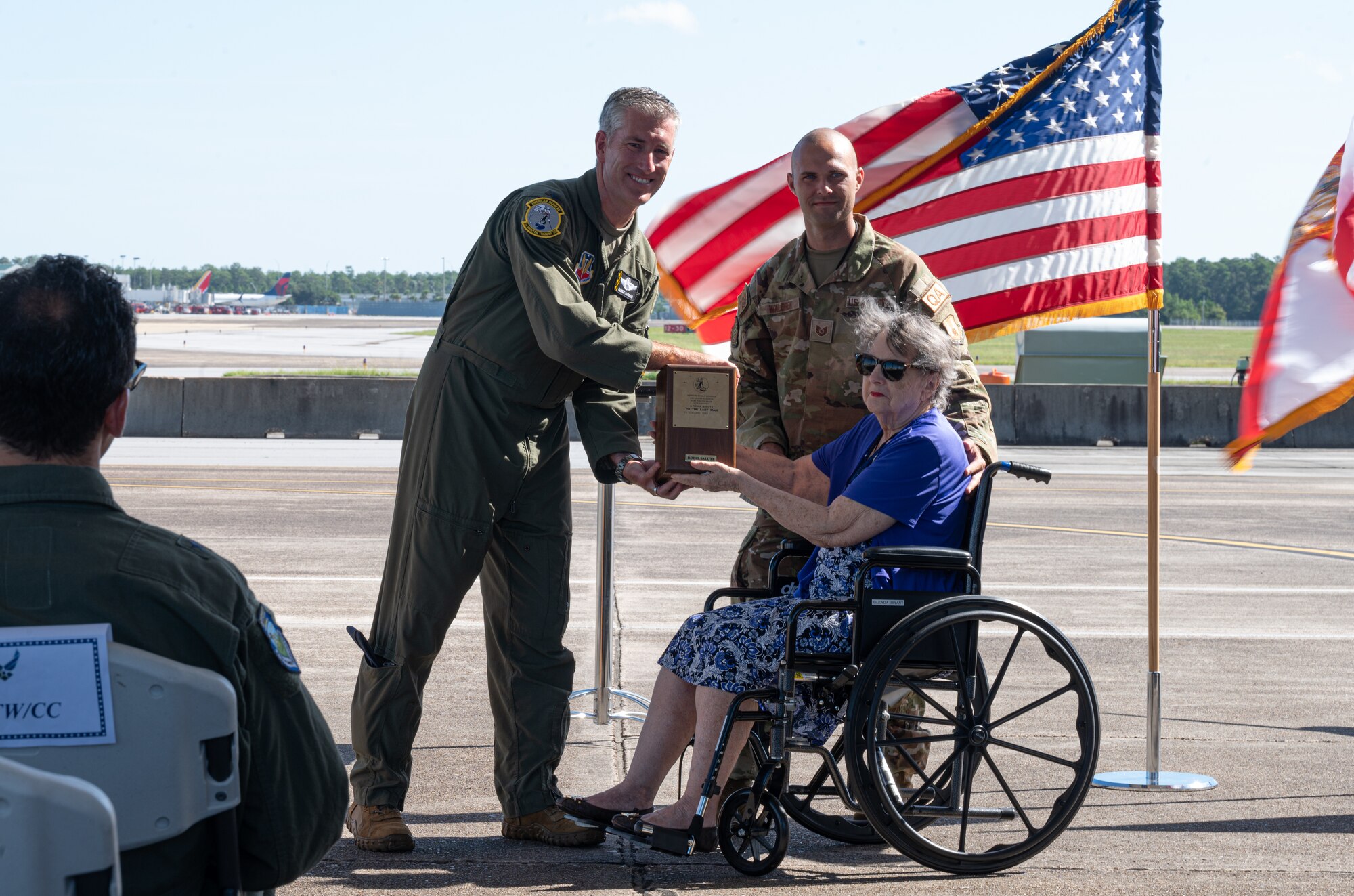 Tyndall's commander accepts a bottle of scotch from a woman in a wheelchair as her grandson stands behind her