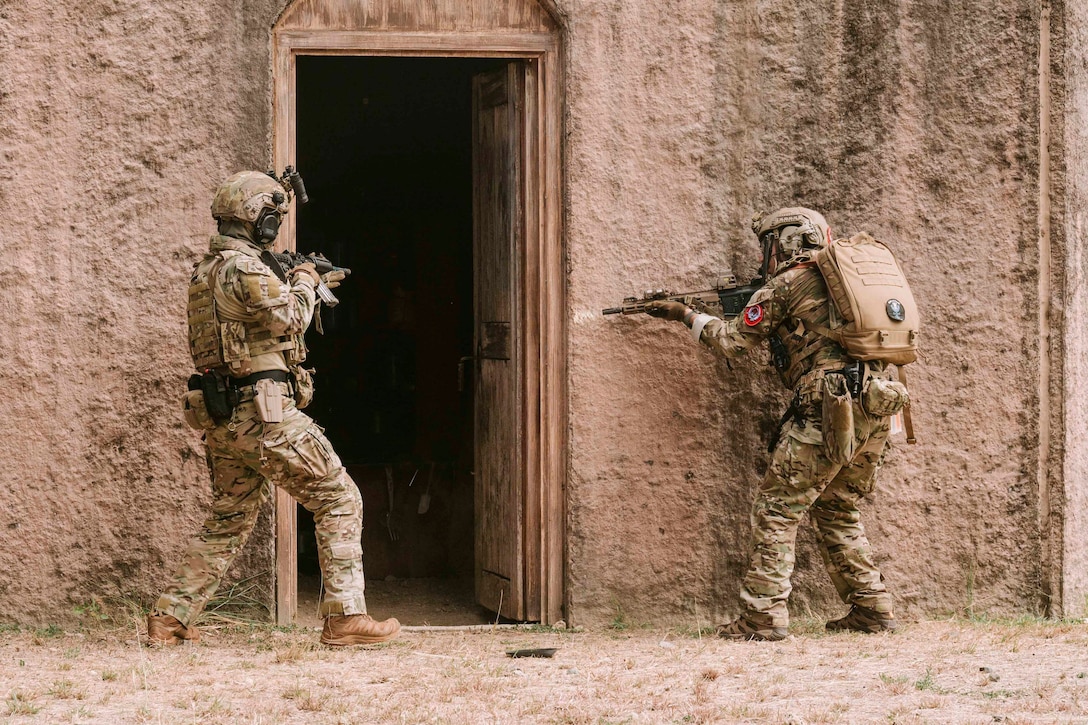 Two service members stand on either side of an open door while holding weapons.