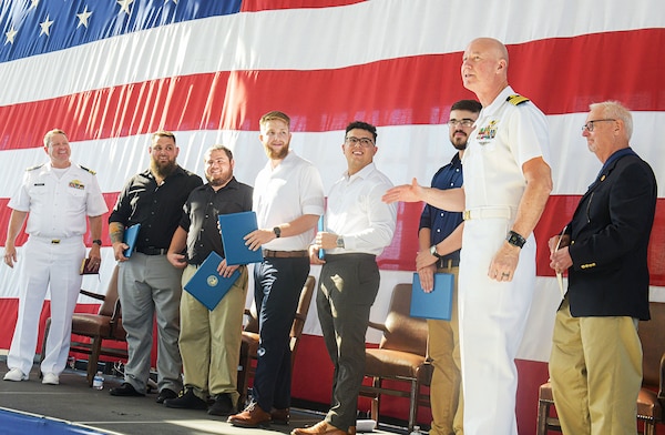 Capt. Grady Duffey, Fleet Readiness Center Southeast’s (FRCSE) commanding officer, asks the audience for another round of applause to congratulate the 2022 apprentice graduates. All five graduating apprentices were in the painting discipline concentration at FRCSE. FRCSE launched the renewed apprenticeship program in 2019, a combination work-study program that provides apprentices with a competitive wage and benefits while learning a journey-level trade. L to R: LCDR Joel DeGraeve, Joshua Lancaster, Andre Giannini, Robert Boehnel, Gilbert Baez, John McCarthy, CAPT Grady Duffey, Mr. Joe Pickens, and Reginald Valentine. (U.S. Navy Photo by Toiete Jackson/Released).