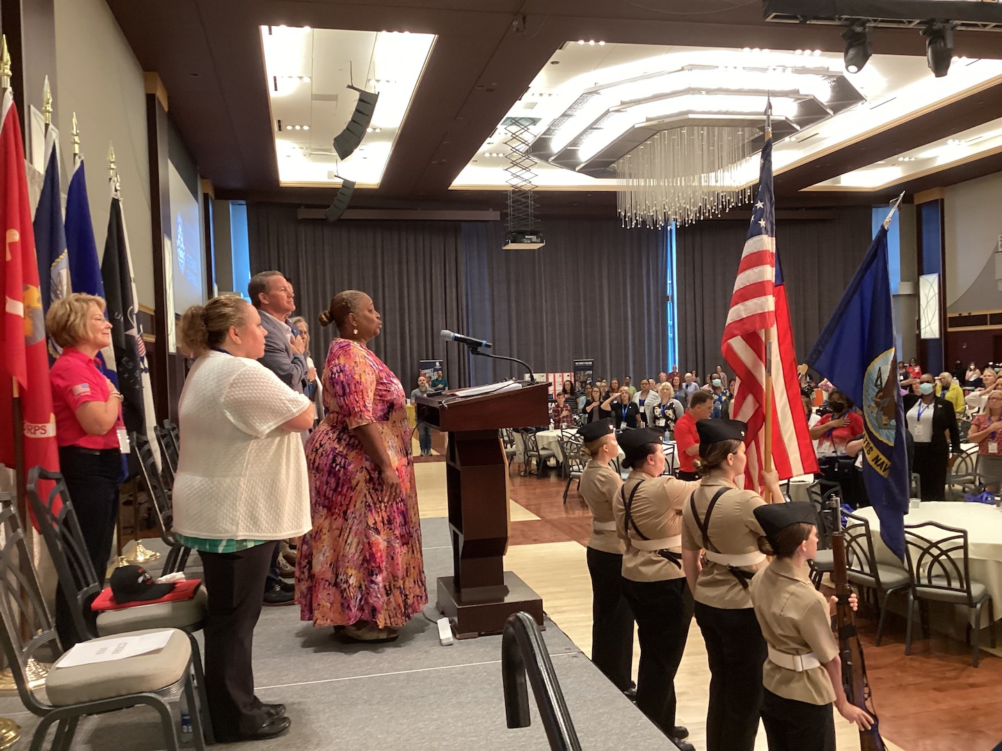 A group of people both men and women in business dress stand on stage in a large ballroom with hands on their hearts as a heavyset woman in a pink flowered dress sings the National Anthem. In front of her are a color guard of Navy JROTC students in their Navy uniforms. Two hold rifles and two hold flags. One is the US Flag, the other the Navy flag.