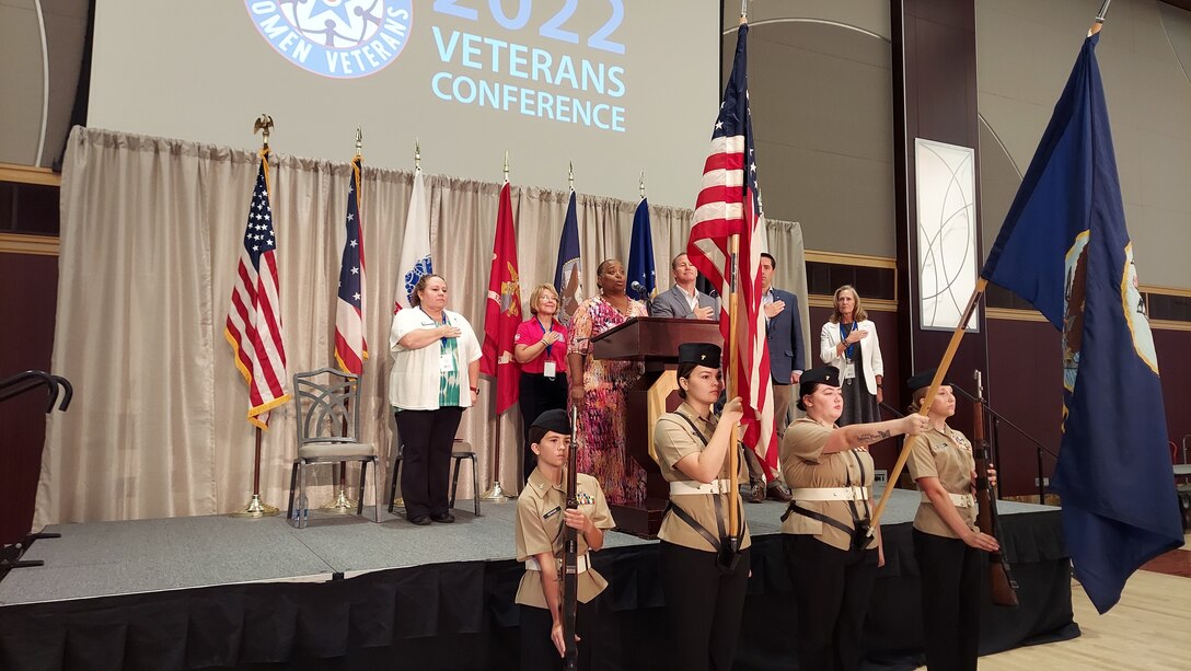 A group of people both men and women in business dress stand on stage in a large ballroom with hands on their hearts as a heavyset woman in a pink flowered dress sings the National Anthem. In front of her are a color guard of Navy JROTC students in their Navy uniforms. Two hold rifles and two hold flags. One is the US Flag, the other the Navy flag.