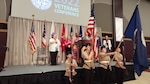 A group of people both men and women in business dress stand on stage in a large ballroom with hands on their hearts as a heavyset woman in a pink flowered dress sings the National Anthem. In front of her are a color guard of Navy JROTC students in their Navy uniforms. Two hold rifles and two hold flags. One is the US Flag, the other the Navy flag.