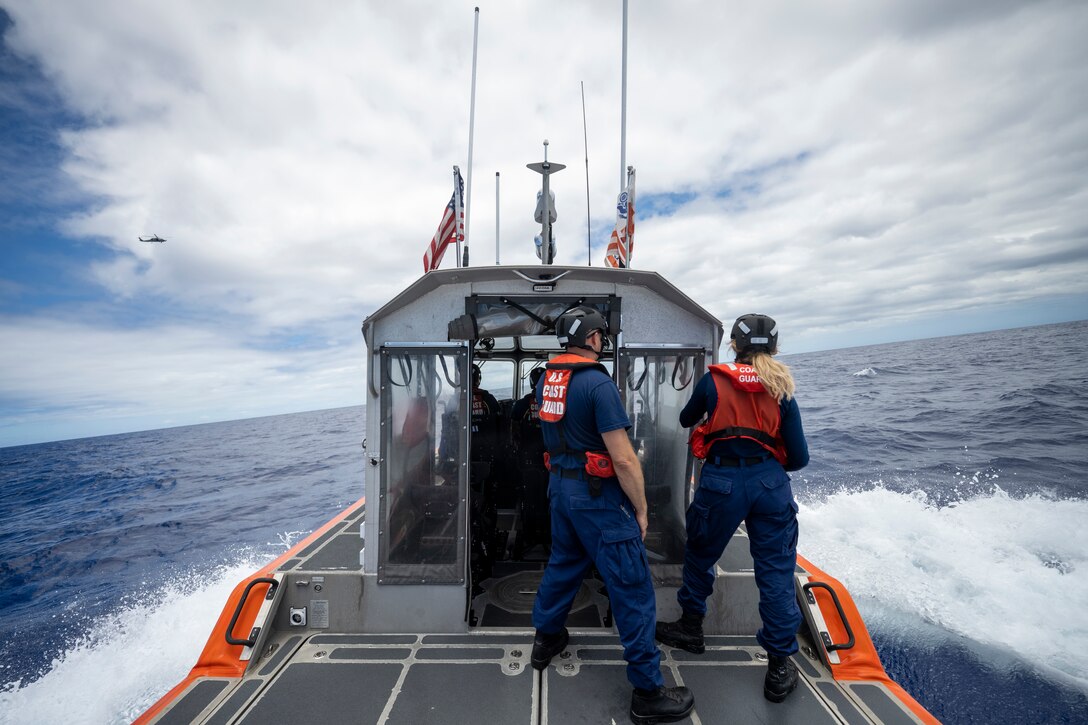 Two Coast Guardsmen stand in the back of a boat moving through the water as a helicopter flies overhead.