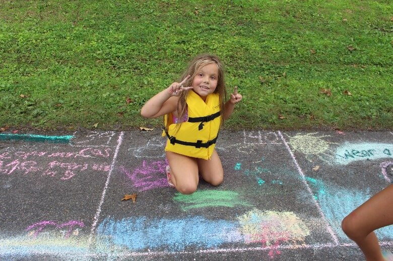 A girl showing the peace sign while in a life jacket coloring with chalk on the sidewalk