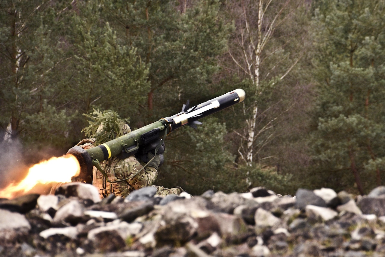 A solider sits in a rocky area with a rocket launcher on his shoulder. Fire is emitted from the rear of the launcher.