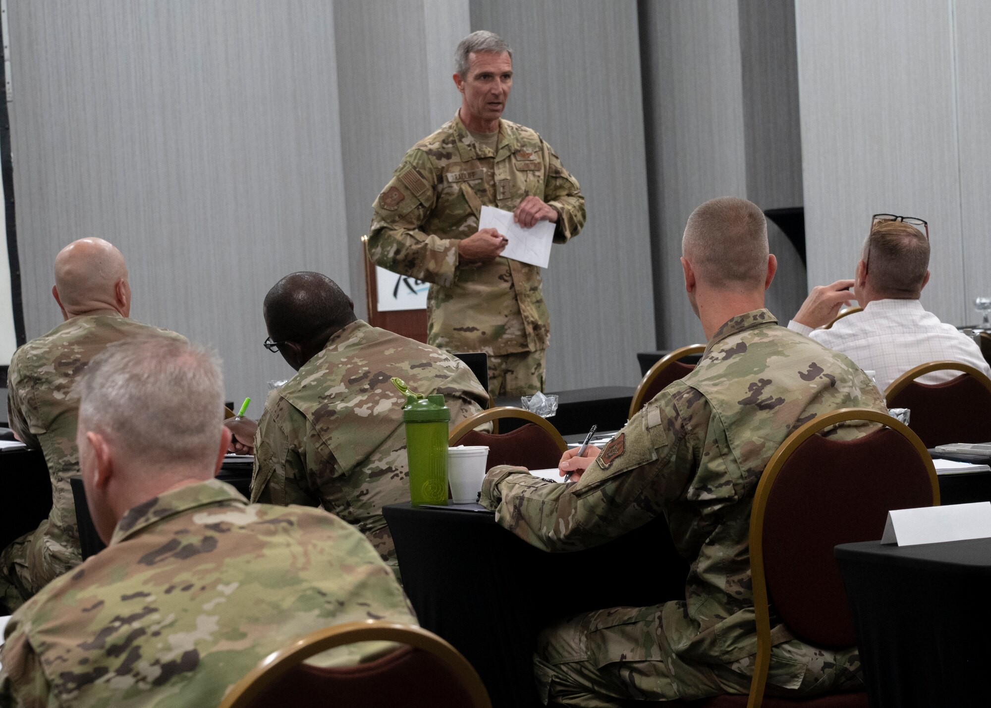 Man in Air Force uniform points to paper while addressing a crowd of 40 people.