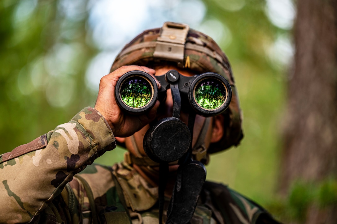 A soldier looks through binoculars in a forest environment.