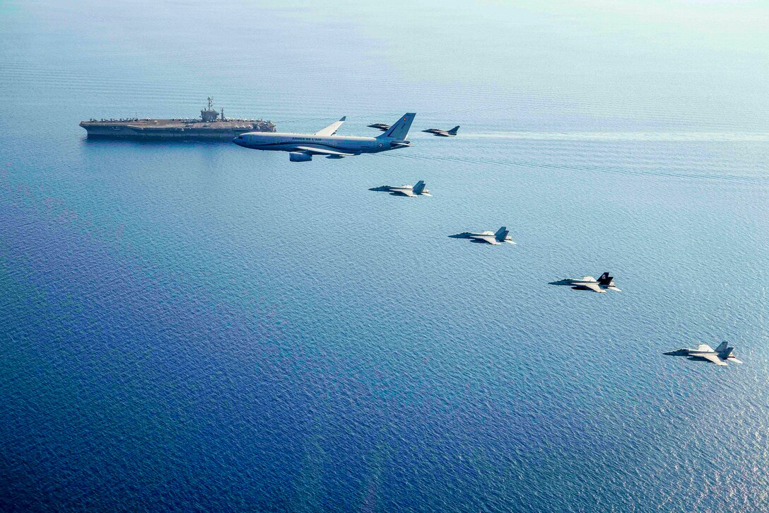 Military aircraft fly in a “V” formation over near a large aircraft carrier in the sea below.