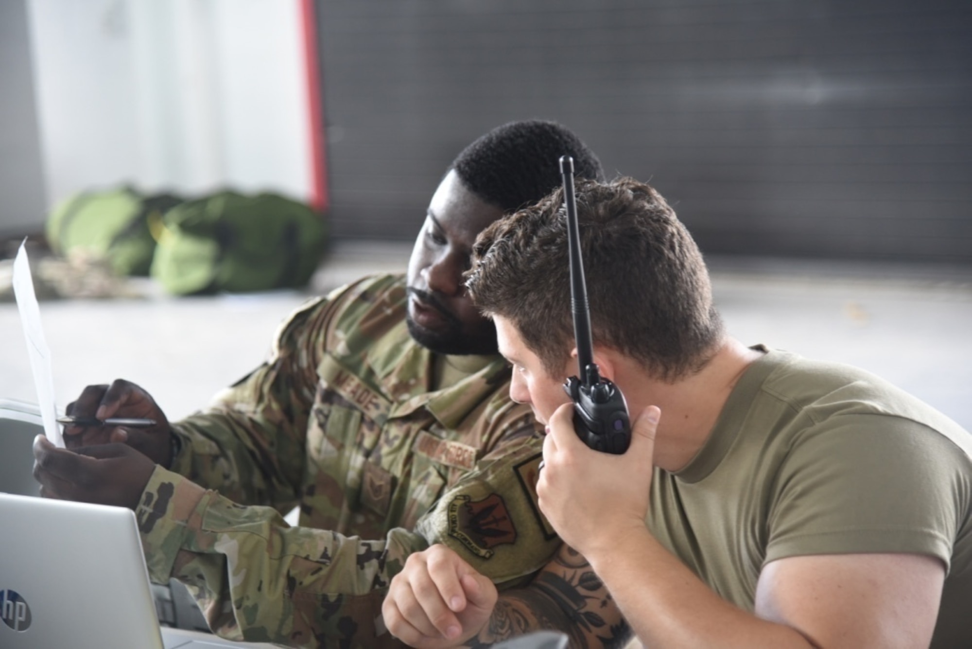 Two men in military uniform sitting at a table, one with a hand-held radio.