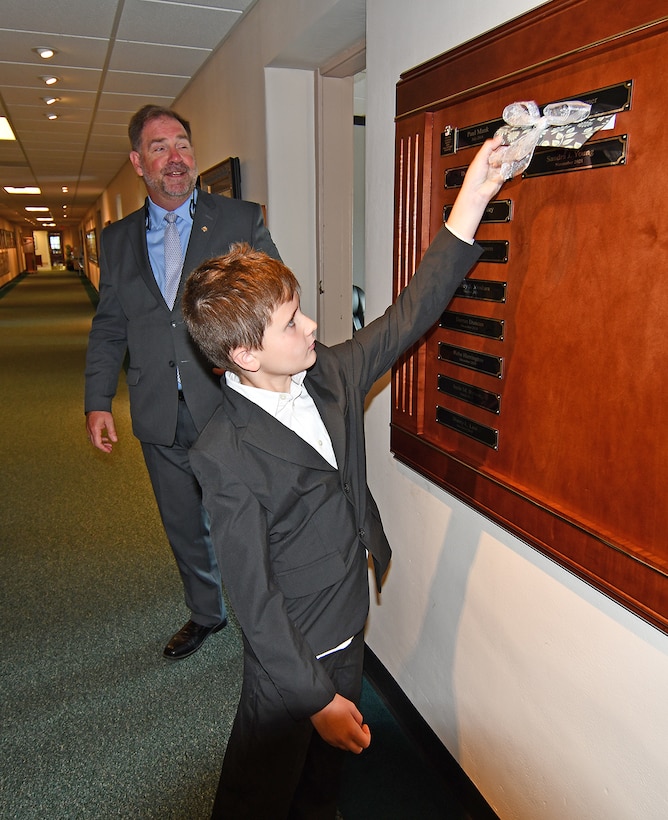 A boy removes a piece of tape from a name plaque on a wall in a hallway.