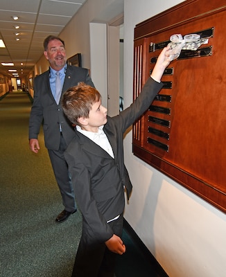 A boy removes a piece of tape from a name plaque on a wall in a hallway.