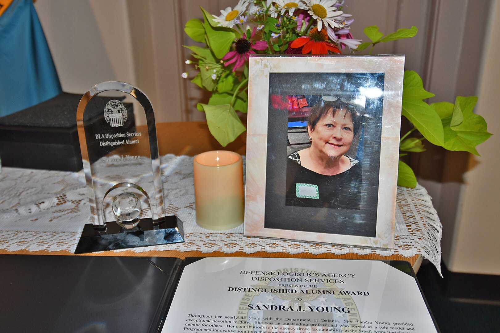 Flowers, a photo, an award citation and a plaque sit on a table with a candle.