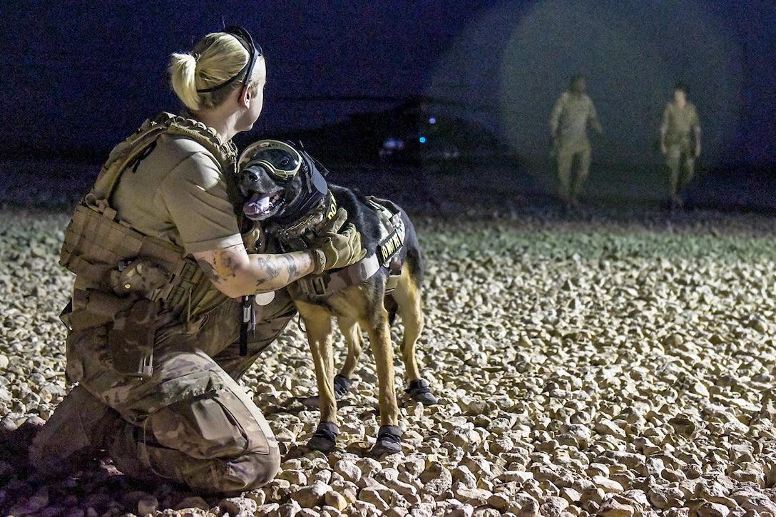 An airman kneels and holds onto a military working dog in booties and goggles near a helicopter at night.