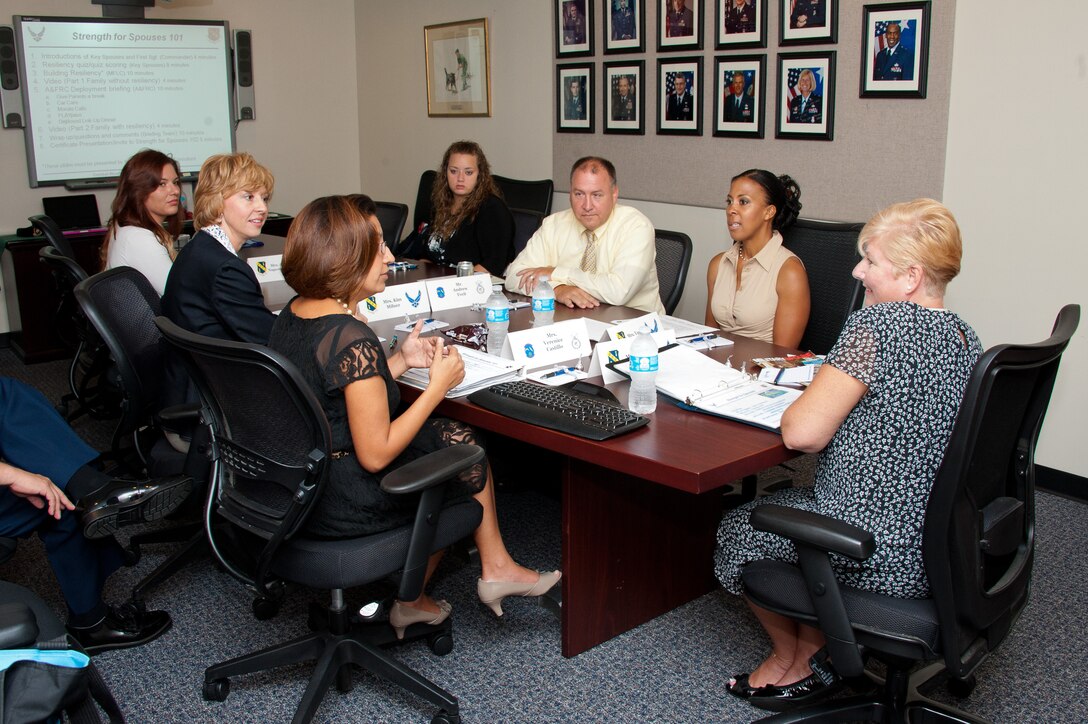 A group of people sit at a conference table, and a presentation with a slide that reads “Strength for Spouses 101” is displayed on a screen.
