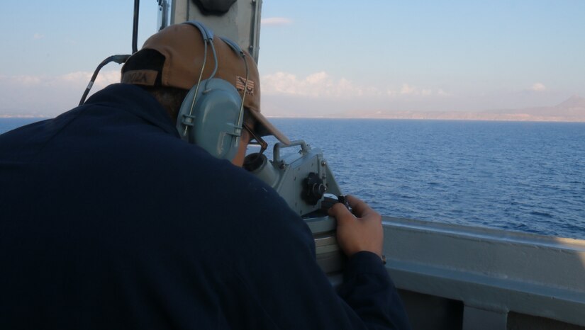 A sailor looks out to sea from behind binoculars aboard a ship.