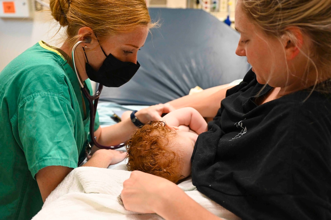 An airman checks vitals on a baby held by a woman.