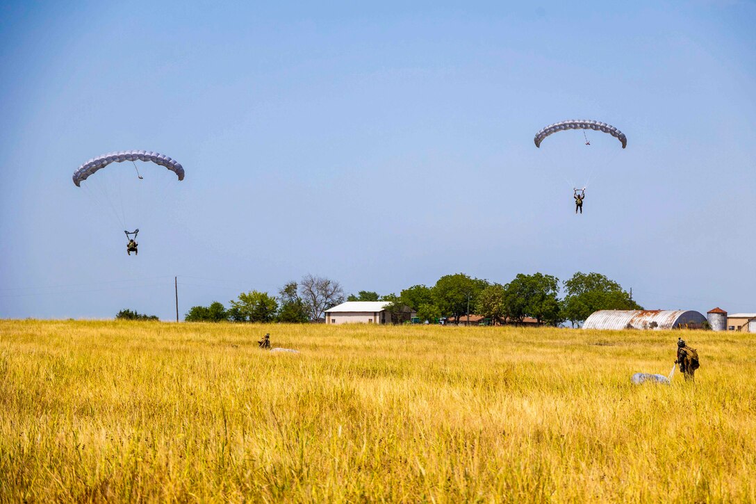 Two Marines descend in the sky wearing parachutes while others walk in a grassy field below.